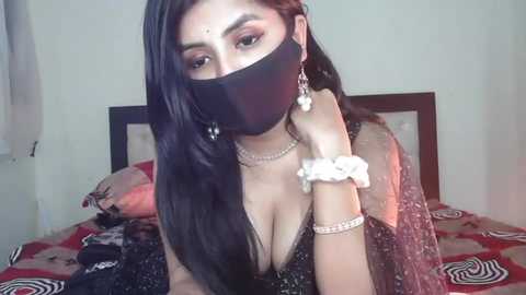 Media: Video of a South Asian woman with long black hair, wearing a black face mask, black dress, and several accessories, sitting on a bed with a patterned blanket in a dimly lit room.