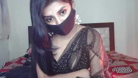 Media: Video of a woman with long black hair, wearing a black sari with floral patterns, a black face mask, and large silver earrings, sitting on a bed with a red and white patterned blanket.