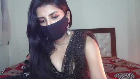 Media: Video of a South Asian woman with long black hair, wearing a black mask, sparkling gold sari, and pearl earrings, sitting on a bed with a patterned red and black bedspread.