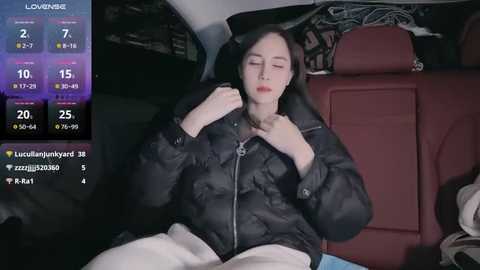 Media: A video of a woman with fair skin and long black hair, wearing a black puffer jacket, asleep in a car. The car's interior is dimly lit, with a red leather seat.