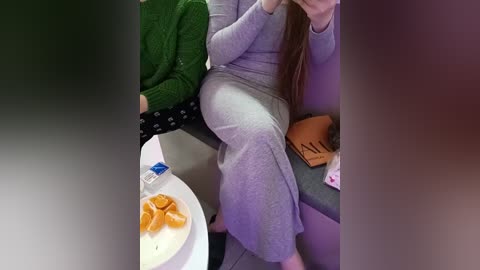 Media: Video of a woman in a gray, loose-fitting outfit, sitting on a purple couch, holding a phone. Her long, straight brown hair drapes over her shoulder. In the background, a green sweater and a brown bag are visible.