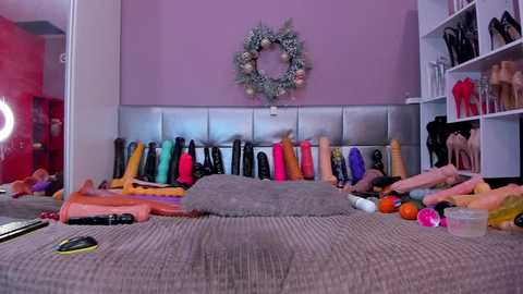 Media: Video of a bedroom with a bed covered in various colored sex toys, a silver headboard, and a pastel purple wall adorned with a decorative wreath.