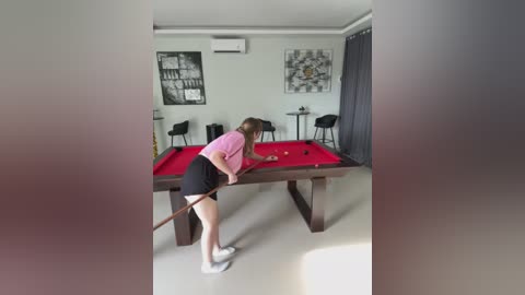 Media: Video of a young woman in a pink shirt and black shorts playing pool in a modern, minimalist room with white walls, dark curtains, and black chairs.