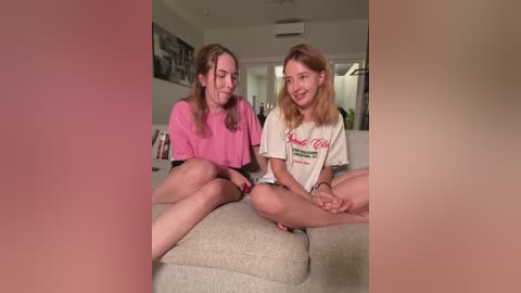 Media: Video of two young women with fair skin and light brown hair, one in a pink t-shirt, the other in a white shirt, sitting on a beige couch in a modern living room.