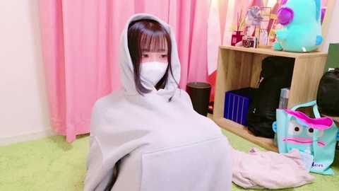 Video of an East Asian woman with long black hair, wearing a white mask, gray hoodie, and pink scarf, sitting on a green carpet, surrounded by stuffed animals and a blue bag.