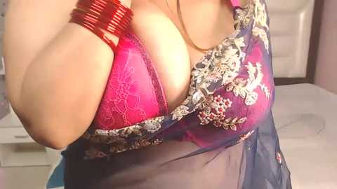 Video of a South Asian woman in a bright pink lace bra with intricate embroidery, under a sheer blue robe. Wearing red bangles, she stands in a modern bedroom with white bedding.