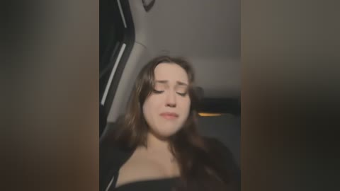 Media: Video of a young woman with long brown hair, wearing a black top, sleeping with eyes closed in the backseat of a car. The background shows the car's interior with a blurred view of the driver's seat.