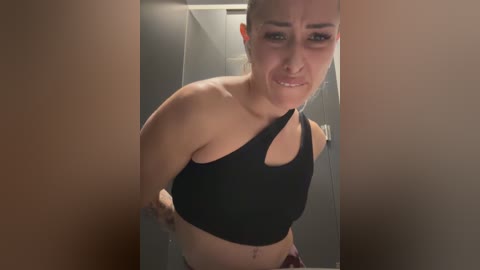 Media: Video of a woman with light skin, medium build, and a black sports bra, standing in a dimly lit hallway, looking upset, with a narrow frame.