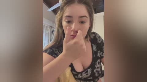 Media: A video of a young woman with light skin, straight brown hair, and wearing a black top with a white pattern, examining her face with her fingers in a casual, indoor setting.