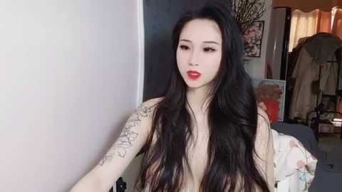 Media: Video of an Asian woman with long black hair, red lipstick, and a tattoo sleeve, standing in a dimly lit room with a bed, clothes, and a mirror in the background.