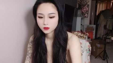 Media: Video of a young East Asian woman with long black hair and fair skin, wearing red lipstick and a sleeveless floral dress, standing in a dimly lit room with floral arrangements, a chair, and a black umbrella.