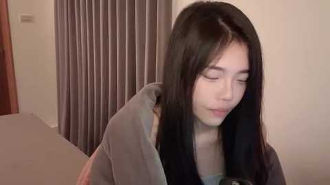 Media: A video of an Asian woman with long, straight black hair, fair skin, and a slender physique, wearing a grey hoodie, standing in a room with beige walls and grey curtains.