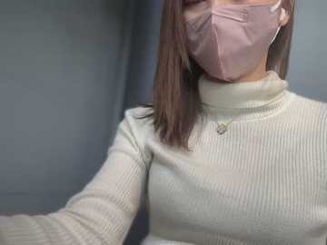 Media: Video of a woman with medium skin tone wearing a beige ribbed sweater, a pink face mask, and silver earrings, standing indoors against a grey wall.