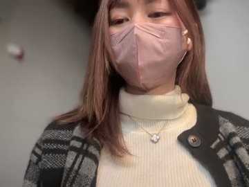 Media: Video of an East Asian woman with light skin, wearing a pink face mask, beige turtleneck sweater, and black-and-white checkered cardigan, with earbuds visible. Background is blurred, indoor setting.