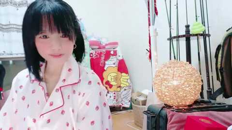 Media: Video of a young Asian woman with short black hair, wearing a white pajama top with red lipstick patterns, standing in a cluttered bedroom with a wooden floor, a red suitcase, and a large decorative lamp.