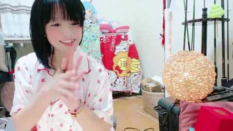 Media: A video of a young Asian woman with straight black hair, wearing a white pajama top with red hearts, smiling and making a peace sign. Background shows a cluttered room with colorful clothes, a red bag, and a decorative lamp.