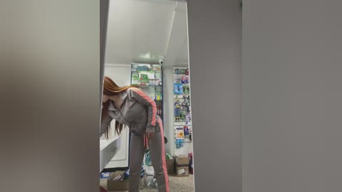 Media: Video of a woman in a gray jumpsuit, bending over, reflected in a mirror. Background shows a cluttered room with shelves of various items.