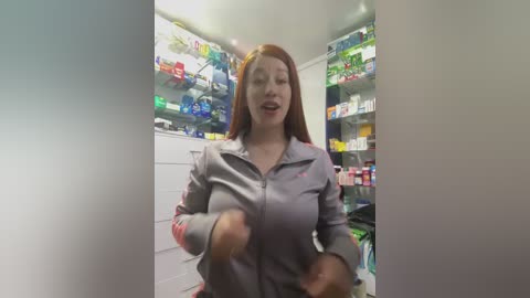 Video of a woman with long, straight red hair in a gray jacket, excitedly dancing in a brightly lit pharmacy. Shelves behind her are stocked with colorful medicine bottles.