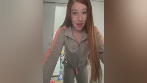 Media: Video of a young woman with long, straight auburn hair, wearing a grey tracksuit with pink accents, leaning forward with a playful expression. The background shows a white door and a messy room with toys.