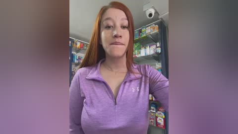 A video of a fair-skinned, red-haired woman with medium-sized breasts wearing a lavender zip-up shirt in a brightly lit, messy grocery store aisle.