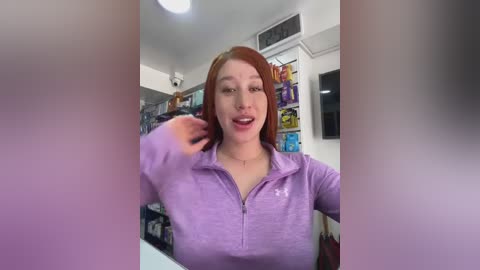 Media: A video of a smiling woman with red hair, wearing a purple zip-up jacket, standing in a brightly lit convenience store with shelves of snacks and drinks in the background.