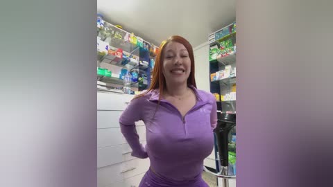 Media: Video of a smiling, fair-skinned woman with red hair, wearing a purple zipped-up top, standing in a brightly lit, cluttered pharmacy aisle.