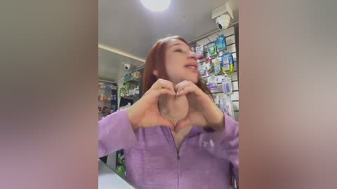 Media: Video of a young Asian woman with straight, shoulder-length brown hair, smiling and forming a heart with her hands in a pharmacy. She wears a lavender zip-up jacket. Background shows shelves of medication.