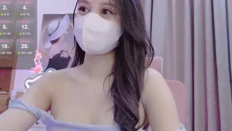 Media: Video of an East Asian woman with long black hair, wearing a white face mask, sitting in a pink chair. Background includes a digital clock and a pink floral painting.