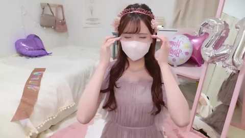 Media: A young Asian woman with long brown hair, wearing a light purple dress, white mask, and headband, adjusts her glasses in a pastel-pink bedroom with heart-shaped decorations, a bed, and balloons.