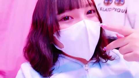 Media: Video of an East Asian woman with straight dark brown hair and bangs, wearing a white mask, light blue jacket, and pink nails, against a pink backdrop, smiling.