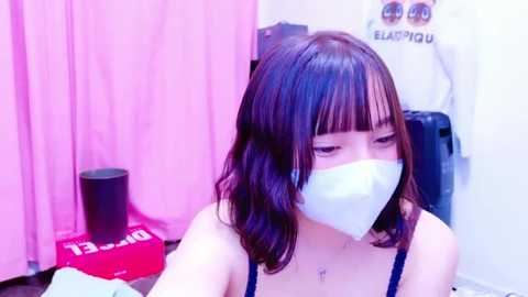 Media: Video of an Asian woman with shoulder-length dark hair, wearing a blue mask, pink and white background, holding a selfie stick, a black speaker, and a white jacket visible.