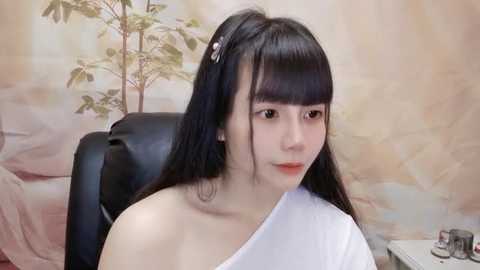 Media: Video of a young East Asian woman with straight black hair, wearing a white off-shoulder top, seated in a black office chair, against a pastel floral wallpaper backdrop.