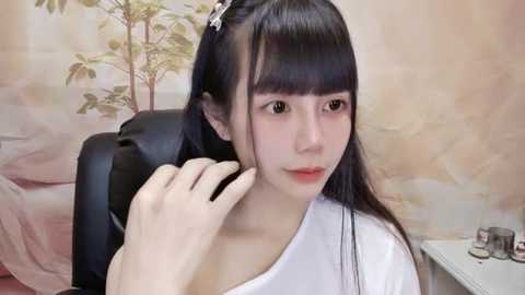 Media: Video of a young Asian woman with long, straight black hair and bangs, wearing a white top, sitting in a black office chair. She has a serene expression and is adjusting her hair. Background features a floral wallpaper and a small table with a cup.