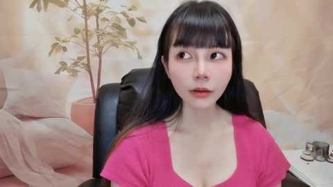 Media: Video of an East Asian woman with straight black hair and pale skin, wearing a pink T-shirt, sitting in a black office chair against a beige, floral-patterned wall with a potted plant.