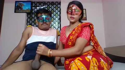 Media: Video of an Indian man and woman in traditional attire, wearing colorful masks, sitting on a bed.