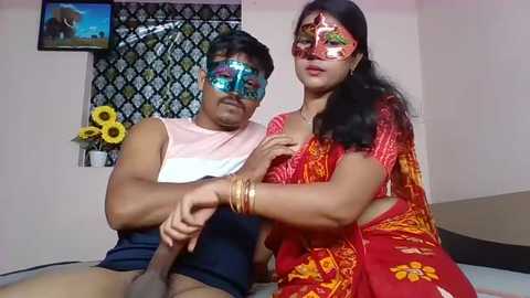 Media: Video of a muscular Indian man with a black mask, wearing a white tank top and shorts, and a woman in a red sari, also masked, in a bedroom with floral decor and a TV.