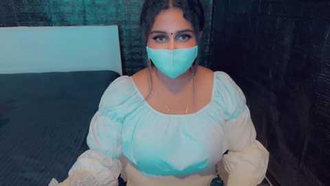 Media: Video of a woman with light brown skin, wearing a turquoise face mask and a white blouse, sitting on a bed in a dimly lit room with dark walls.