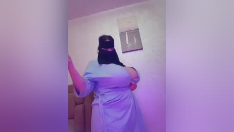 Media: Video of a woman with large breasts, wearing a light blue dress and black face mask, standing in a narrow hallway with beige walls and a framed picture.