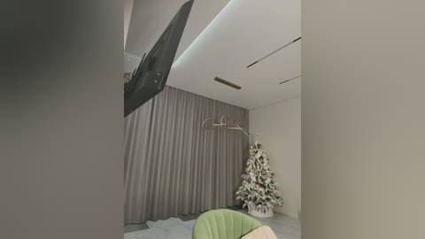 Media: Video of a modern living room with beige curtains, a large TV mounted on the wall, a white Christmas tree adorned with white ornaments, and a light green armchair.