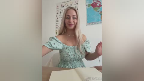 Media: Video of a smiling, fair-skinned blonde woman in a mint green off-shoulder top, seated at a wooden table, holding a book, in a classroom with alphabet and map posters on the wall.