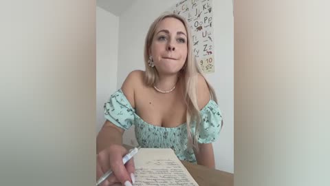 Media: Video of a blonde Caucasian woman with fair skin, medium breasts, and a light green off-shoulder dress, writing in a notebook at a desk, surrounded by alphabet posters on a white wall.