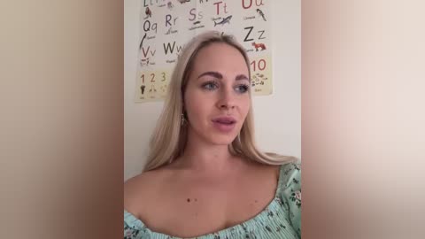 Media: A video of a blonde woman with fair skin, wearing a light blue off-shoulder top, standing against a beige wall adorned with educational posters featuring letters and numbers.