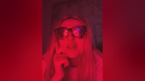 Media: Video of a woman with long blonde hair, wearing oversized black sunglasses and a white top, with a red tinted background, creating a surreal, eerie atmosphere.