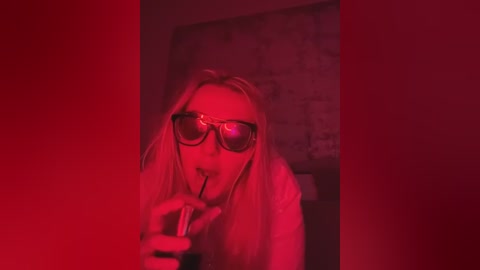 Video of a blonde woman with dark glasses, holding a pen in her mouth, under red lighting. She has a puzzled expression.