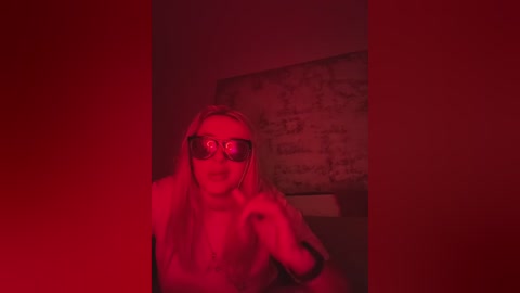 A video of a blonde woman with glasses, wearing a white coat, in a dimly lit room with red lighting, standing in front of a brick wall.