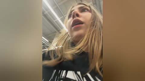 Media: Video of a young, light-skinned woman with shoulder-length blonde hair, wearing a black hoodie with white stripes. She has a slightly open mouth and a contemplative expression, taken from a low angle in an industrial setting with exposed pipes.