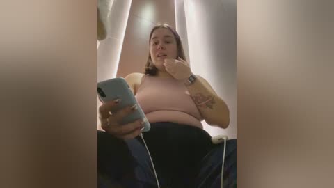 Media: A video of a young woman with medium build, light skin, and shoulder-length brown hair, wearing a pink tank top and dark pants, taking a selfie in a mirror, revealing tattoos on her arm.