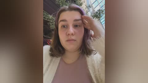 Media: A video of a young woman with shoulder-length, wavy brown hair, wearing a light pink shirt and a white cardigan, standing in a sunlit indoor area with greenery and modern architecture in the background.