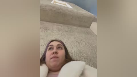 Media: A video of a young woman lying on a beige carpet, looking upwards with a neutral expression. The background shows a white staircase and a blue wall. She has brown hair and wears a light-colored shirt.