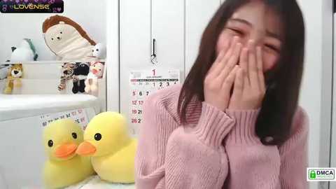 Video of an Asian woman in a pink sweater, laughing with hands covering her face, surrounded by plush toys in a white room with a calendar.